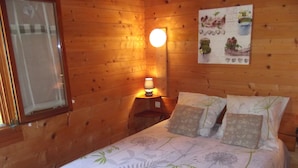 Une pièce en bois avec un lit, une table de chevet, une lampe fixée au mur et une fenêtre.
