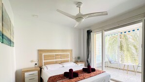 Hotelkamer met een bed, een plafondventilator, een nachtkastje, een raam met uitzicht op palmbomen en een balkon met stoelen.