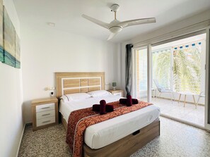 A hotel room with a bed, a ceiling fan, a nightstand, a window with a view of palm trees, and a balcony with chairs.