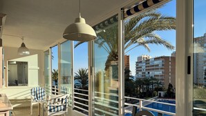 Een balkon met een tafel en stoelen, grote ramen met uitzicht op een zwembad en palmbomen, en een hanglamp.