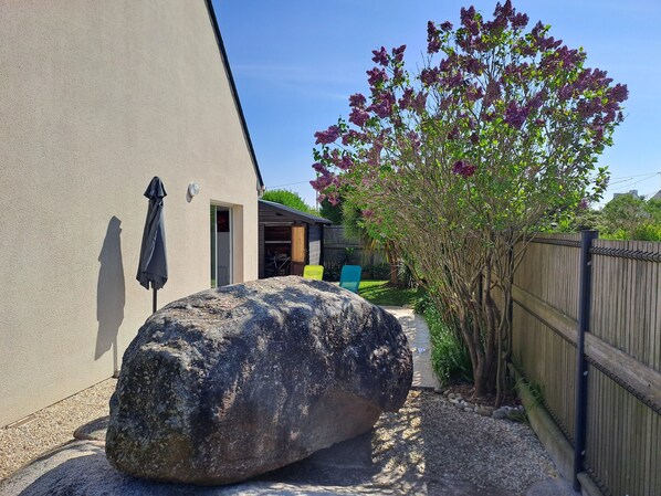 A large rock in a garden area, a wooden fence, and a building with an open door. - From 357€ (ANCV-approved): 3-star single-story house, beachfront, enclosed garden, pets allowed. (Porspoder)