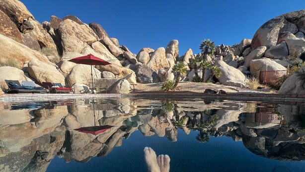 Una piscina cristalina con formaciones rocosas y palmeras, rodeada de sillas de descanso y sombrillas.