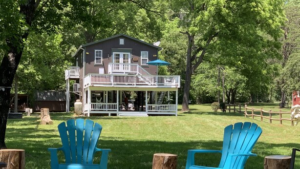 A two-story house with a porch and a deck, surrounded by trees and a fenced area.