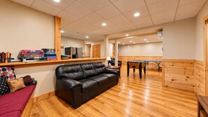 A room with a black leather sofa, wooden flooring, a foosball table, and a shelf with board games. - Grand Ebs View Lodge, Luxury Log Home, Views, Hot Tub, Sauna, EV Charge, 3D Tour (Jay)