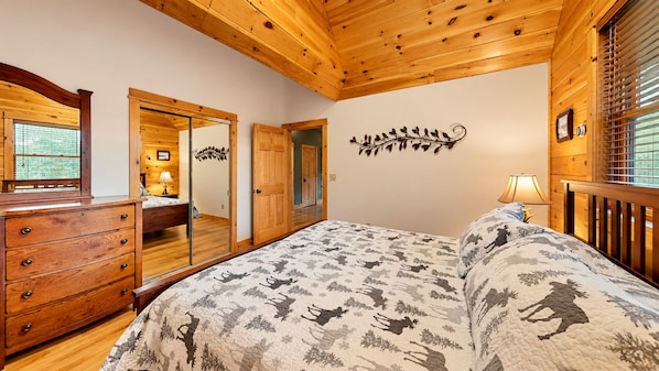 Un dormitorio con techo de madera, una cama con colcha estampada, un aparador, un espejo y un adorno de pared.