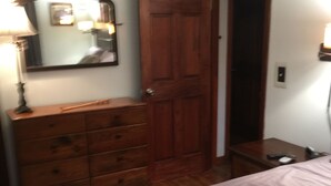A bedroom with a wooden dresser, a bed, a nightstand, a lamp, and a door.