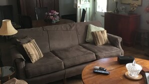 A living room with a brown sofa, a round coffee table, and a dining table. There is a wooden cabinet and a lamp on the table.