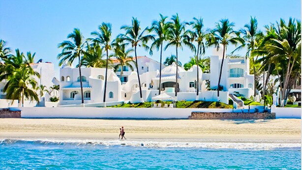 Un hotel frente al mar con edificios blancos, palmeras y una playa.