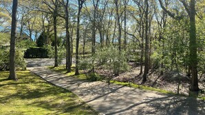 A paved pathway through a wooded area with trees and a house in the background. - Bright and Spacious Summer Home Steps from the Beach (Shelter Island)