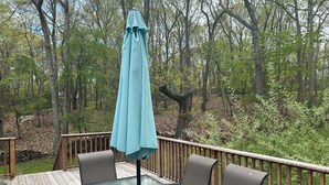 A deck with a glass table and chairs, surrounded by a wooden deck railing, overlooking a wooded area. - Bright and Spacious Summer Home Steps from the Beach (Shelter Island)
