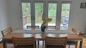 A dining room with a wooden table, four chairs, and a vase of flowers. There are three glass doors leading to an outdoor patio area with a wooden deck and chairs. - Bright and Spacious Summer Home Steps from the Beach (Shelter Island)