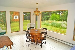 Enjoy meals in this dining area with a round table and four chairs, surrounded by large windows offering a view of the lush garden and brick structure.
