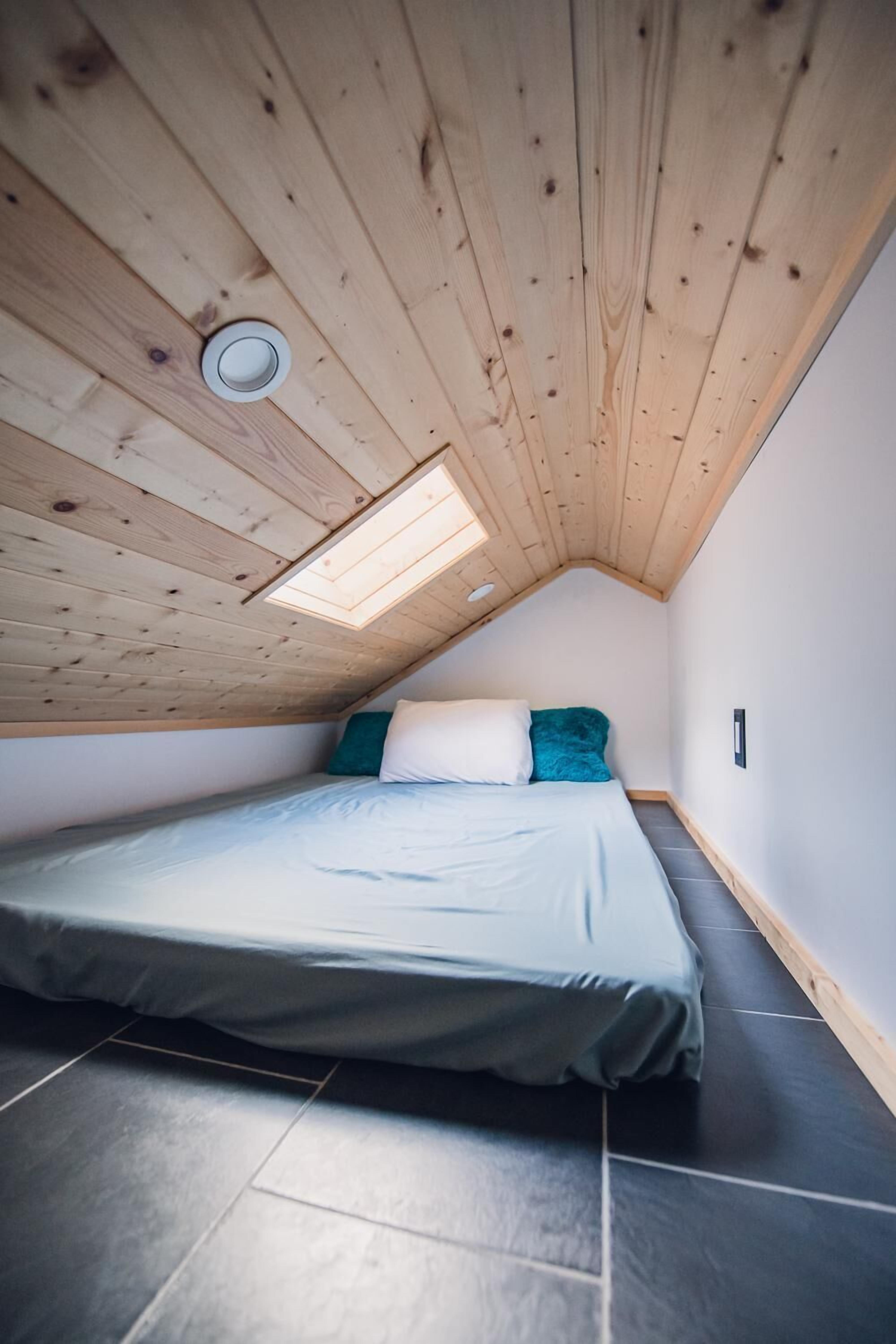 Une petite pièce avec un toit en pente, un lit, un puits de lumière et un luminaire fixé au mur.