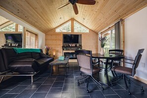 Un salon chaleureux avec un canapé en cuir noir, une table basse et une table à manger avec des chaises. Un téléviseur est fixé au mur et un ventilateur de plafond est installé.