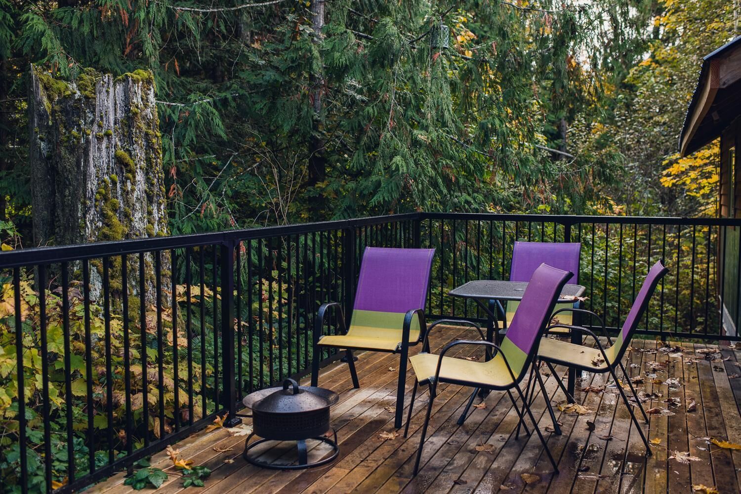 Una terraza de madera con barandilla de metal, rodeada de un bosque frondoso. Hay tres sillas y una mesita.