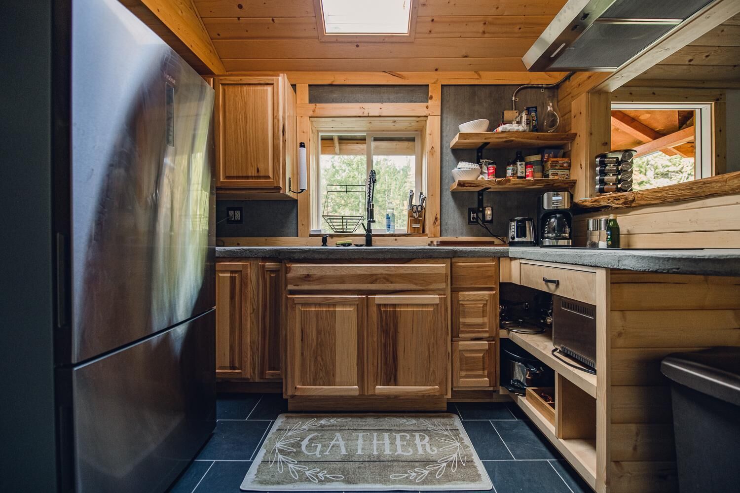 A compact kitchen with wooden cabinets, a stainless steel refrigerator, and a window with a view of greenery.