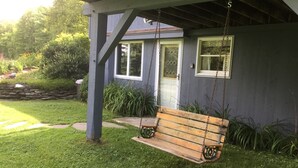 A wooden swing bench under a porch with a stone wall and a house in the background. - Meadows Mountains and more  (Waterbury)