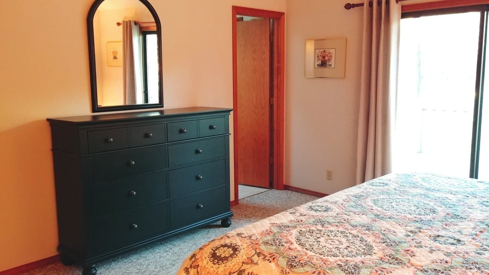 Una camera da letto con una cassettiera scura, un letto con copriletto decorato, uno specchio, una porta e un quadro appeso al muro.
