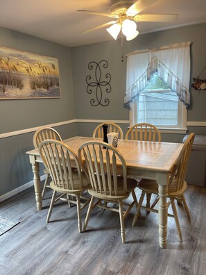 A dining room with a wooden table and chairs, a ceiling fan, a painting, and a window with curtains.