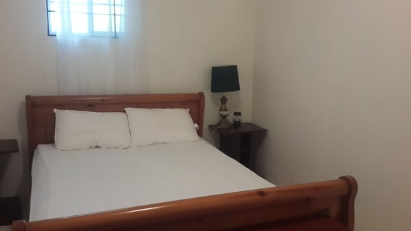 A neatly made bed with white linens and two pillows, a wooden headboard, a bedside table with a lamp, and a window with curtains.
