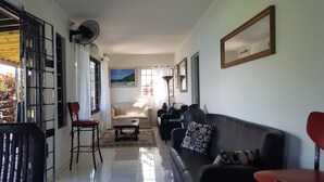 A living room with a sofa, a chair, a table, and a floor lamp. There is a painting on the wall and a window with curtains. - Ridge Bay Chateau (Port Antonio)