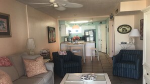 Un salon avec un canapé, deux fauteuils, une table basse et, au fond, une cuisine.