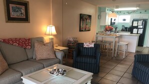 Un salon avec un canapé, un fauteuil et une table basse. Au fond, on aperçoit un coin cuisine avec une table à manger et des chaises.