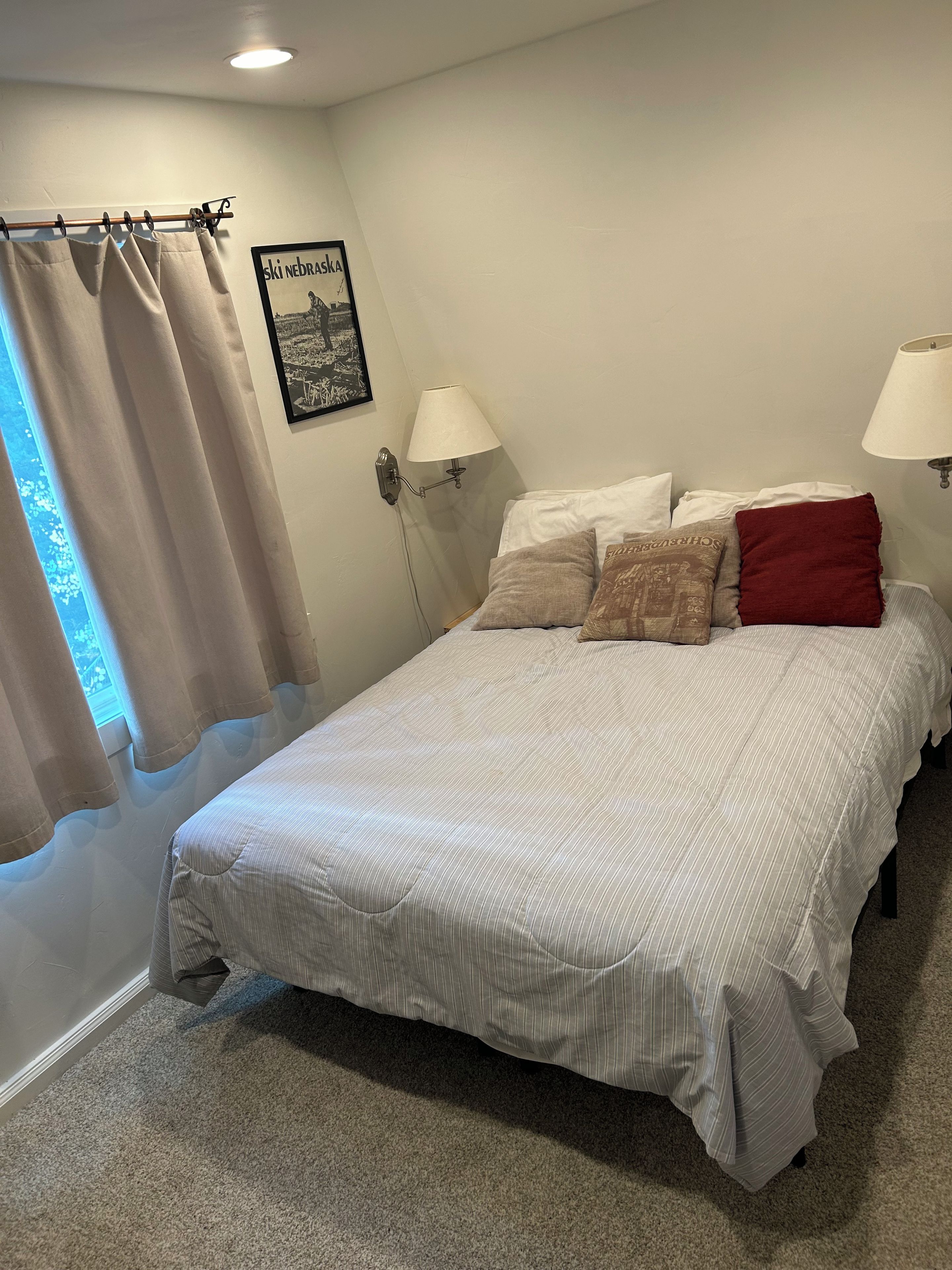 Une chambre à coucher avec un lit, des rideaux, une lampe fixée au mur et un tableau encadré.