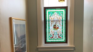 Een gang met een glas-in-loodraam, een lijst met een afbeelding aan de muur, en een houten bankje.