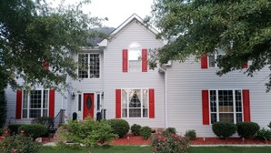 Una casa de dos pisos con contraventanas rojas, un porche delantero y un jardín bien cuidado con acolchado rojo y diversas plantas.