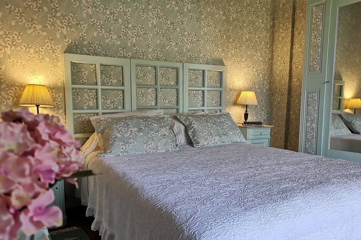 A bedroom with floral wallpaper, a bed with a white bedspread, two bedside lamps, and a mirror on the wall.