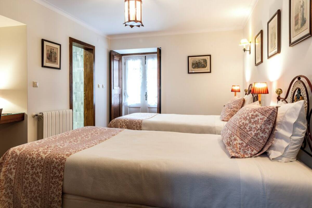 A hotel room with two beds, a wooden door, a radiator, framed pictures on the walls, and a lamp on the nightstand.