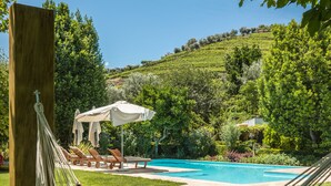 Uma rede, um livro, uma piscina e um jardim com espreguiçadeiras e guarda-sóis.
