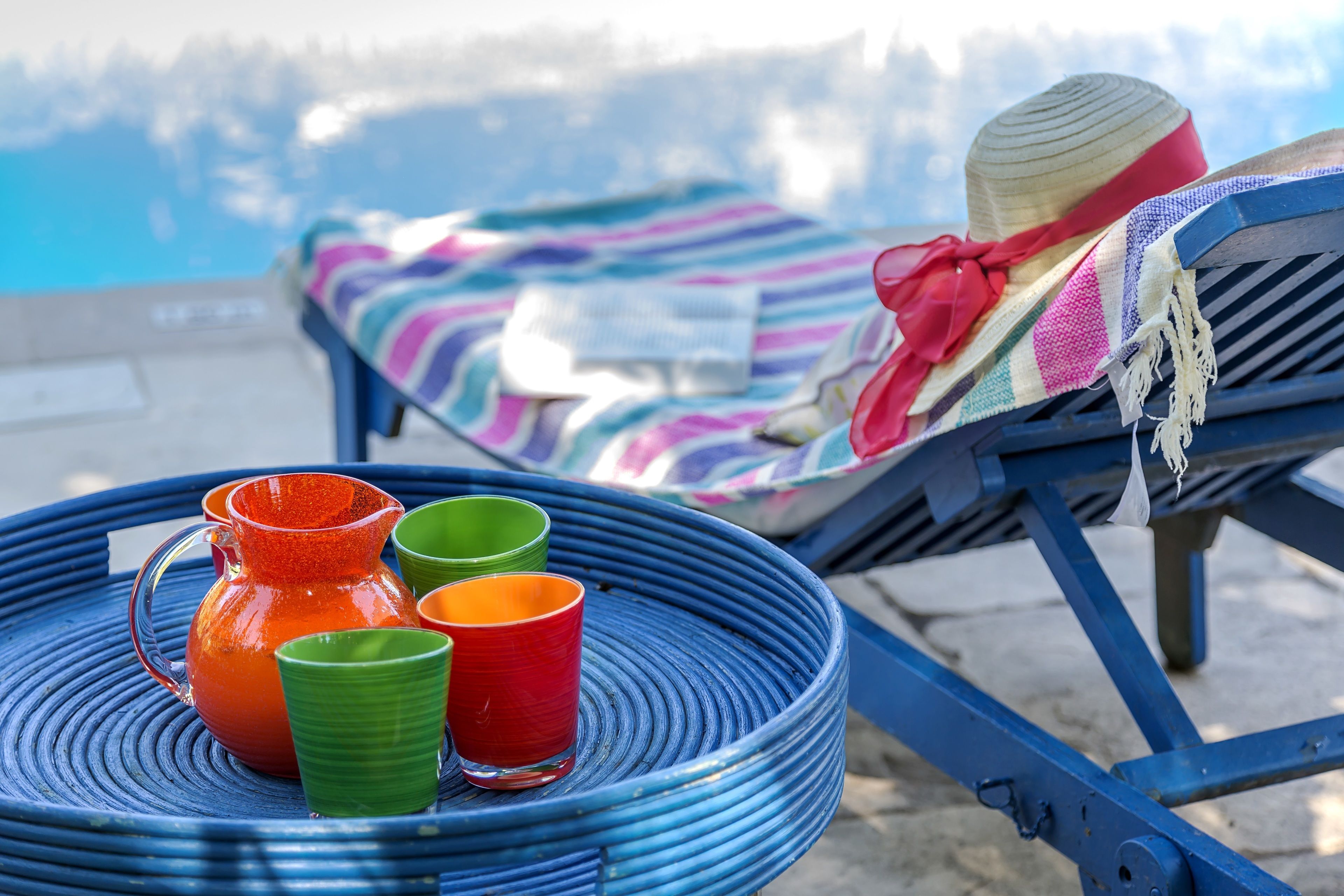 Une chaise longue installée au bord de la piscine, avec une couverture à rayures et un chapeau, une table en osier bleue ornée de tasses colorées et d’une carafe, et une vue dégagée sur la piscine en arrière-plan.