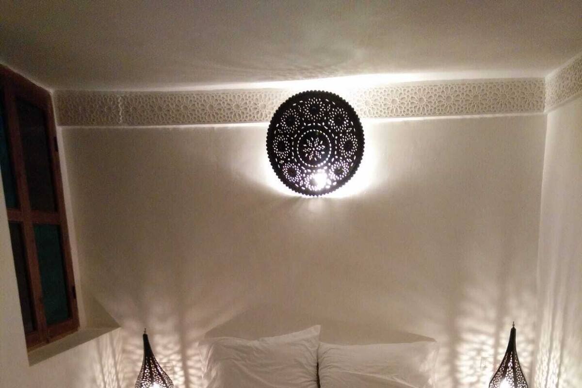 Un dormitorio con una cama grande, dos mesitas de noche, una lámpara decorativa en el techo y una ventana con cortinas.