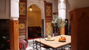 Ein traditionelles marokkanisches Interieur mit aufwendigen Bögen, einem Holztisch mit einem gewebten Korb voller Orangen und einem gemusterten Boden.