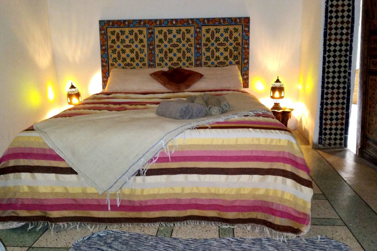Una cama con colcha a rayas, almohadas y una manta. Hay dos lámparas de noche y un panel decorativo en la pared sobre la cama.