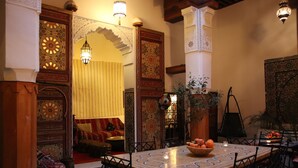 Ein traditionelles marokkanisches Interieur mit einem mit Fliesen ausgelegten Essbereich, verzierten Türen und einer Sitzecke mit Kissen.