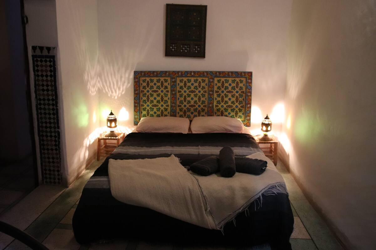 Un dormitorio con cama, dos mesitas de noche con lámparas, cabecero estampado y una obra de arte en la pared.