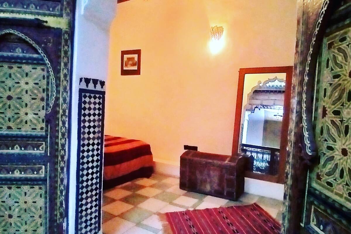 Una habitación con piso a cuadros, un tapete rojo, un banco de madera, una mesita y un espejo en la pared.