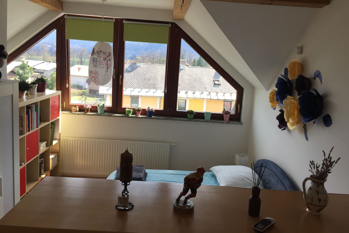 Um quarto com teto inclinado, uma janela grande com vista para casas, uma estante, uma mesinha com uma figurinha e um vaso na parede com flores.