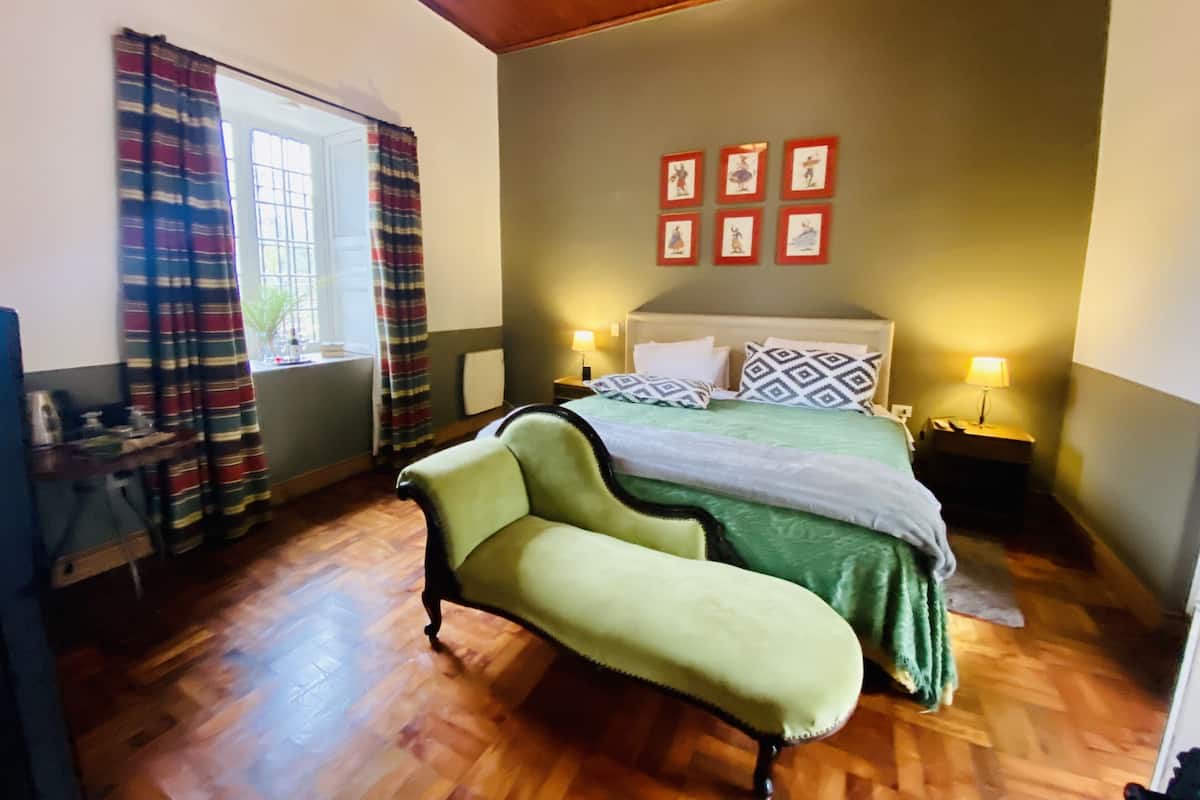 Un dormitorio con cama, una banca, una ventana con cortinas y cuadros en la pared.
