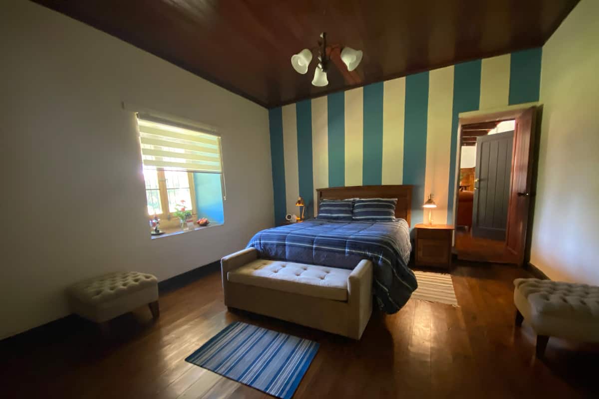 Un dormitorio con techo de madera, una cama con colcha a rayas, una ventana con persianas, una mesita de noche con lámpara y una puerta que da a otra habitación.