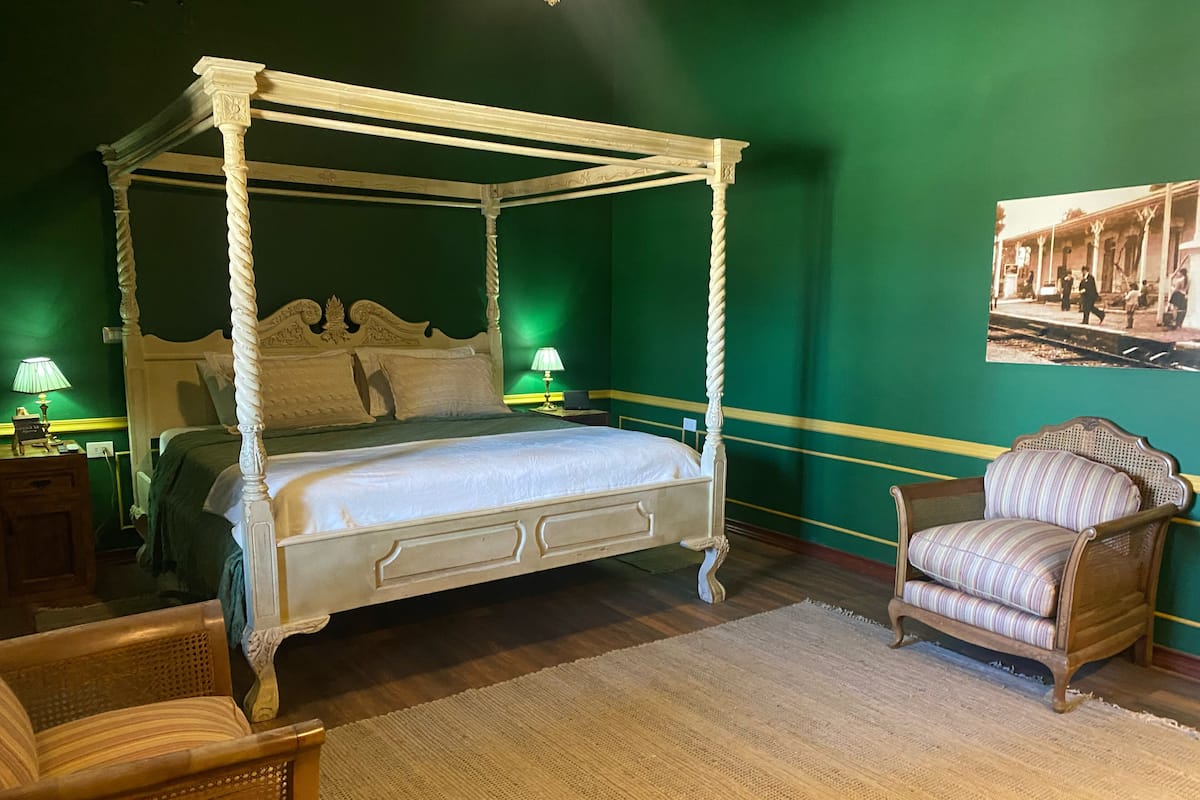 Una cama con dosel sobre cuatro columnas, con estructura de madera y ropa de cama blanca, ubicada en una habitación con paredes verdes y piso de madera.