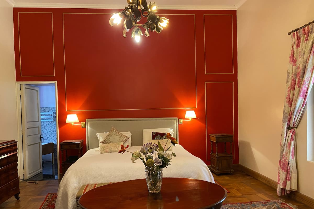 Un dormitorio con una cama grande, dos lámparas de noche, una araña, una mesa de madera con un florero y un tapete con estampado floral.