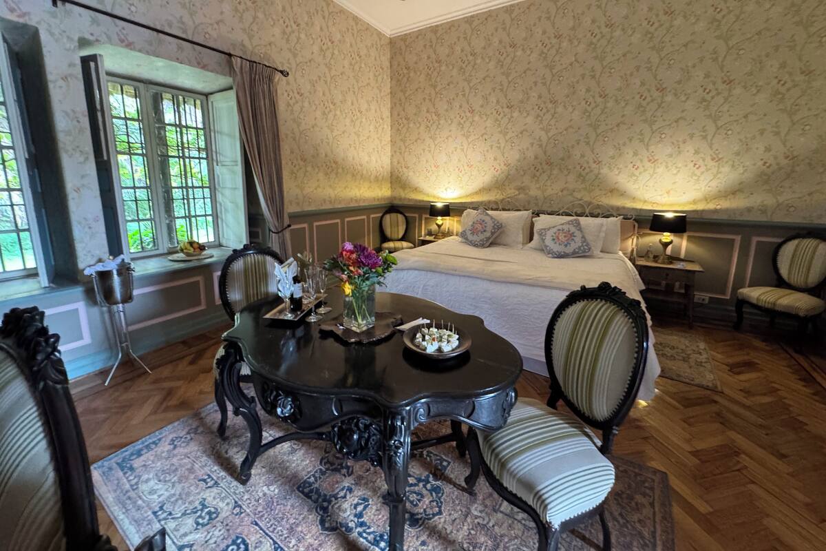 Un dormitorio con una cama grande, una mesa de comedor con sillas, una araña y una ventana con cortinas.