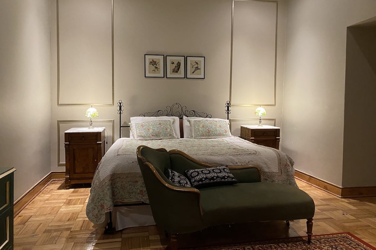 Un dormitorio con una cama grande, dos mesitas de noche, un sillón verde y suelo de madera.