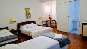 Une chambre d’hôtel avec deux lits simples, une petite table avec des chaises, un tableau au mur et une salle de bain visible à travers une porte ouverte.
