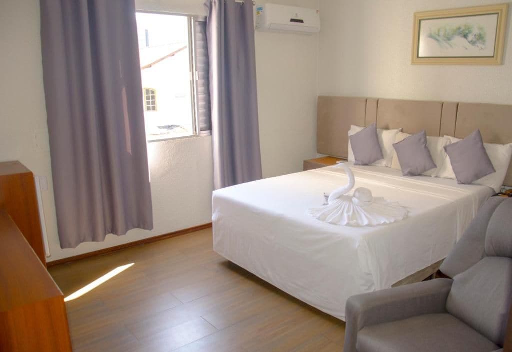 A hotel room with a bed, a nightstand, a chair, a window with curtains, and an air conditioning unit.