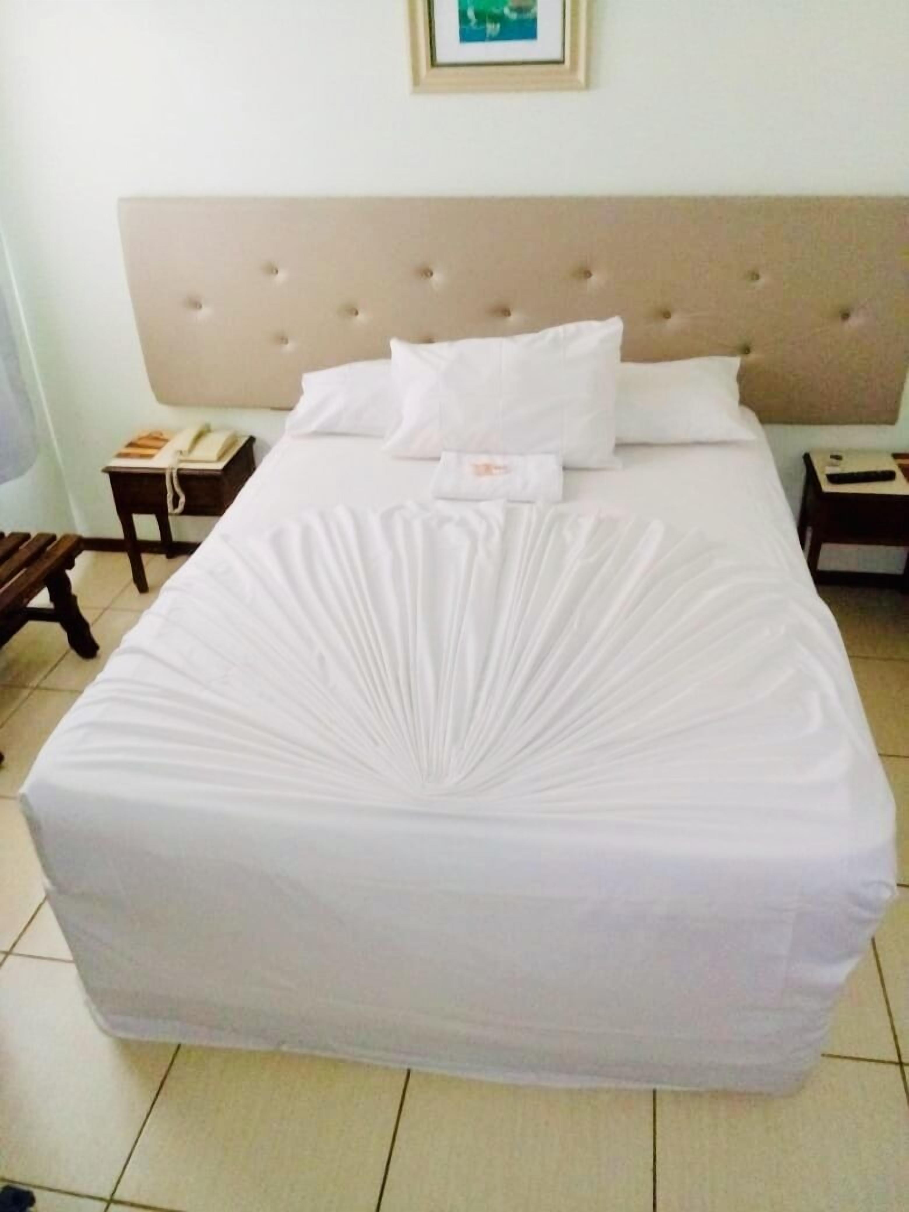 Uma cama bem arrumada com cabeceira estofada, roupa de cama branca e uma pequena mesa de cabeceira com um telefone e um livro.
